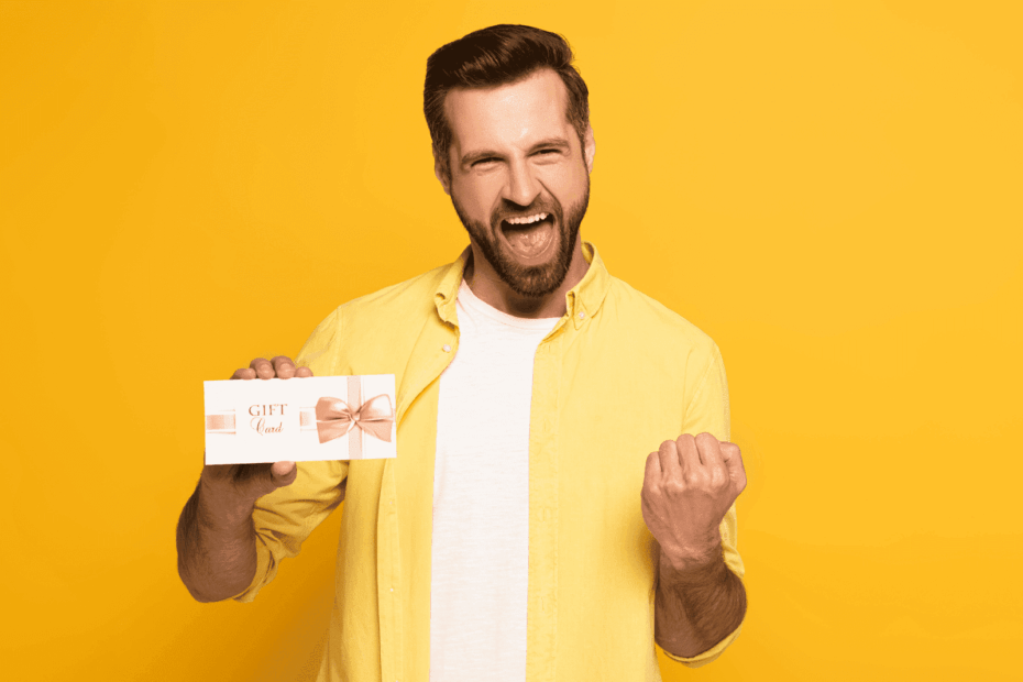 A cheerful man in a yellow shirt holding a white gift card with a pink bow against a bright yellow background