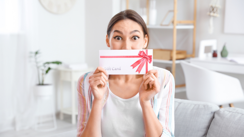 A woman looks surprised and delighted as she holds up a gift card envelope with a red ribbon.
