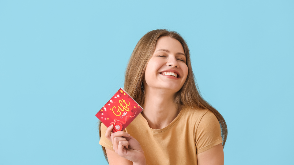A smiling woman joyfully holds up a festive red gift card against a blue background.
