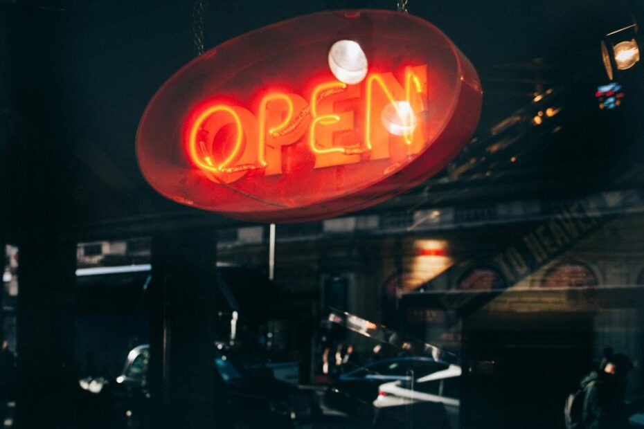 Bright red neon “OPEN” sign hanging in a restaurant’s window with city street reflections in the glass.