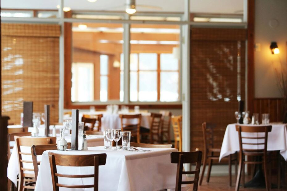 Empty restaurant with wooden chairs and white tablecloths, set for dining with glassware and utensils.