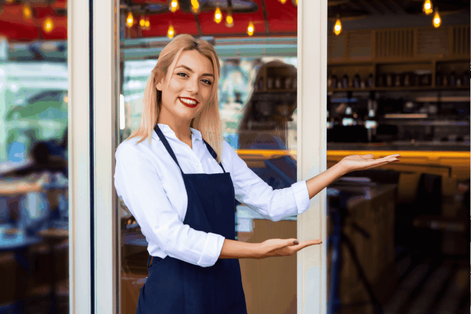 Smiling waitress in a white shirt and dark apron stands at a restaurant entrance, gesturing with open hands to welcome guests inside