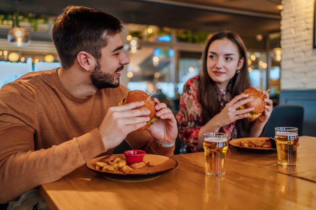 A man and a woman are seated across from each other at a wooden restaurant table, while holding burgers