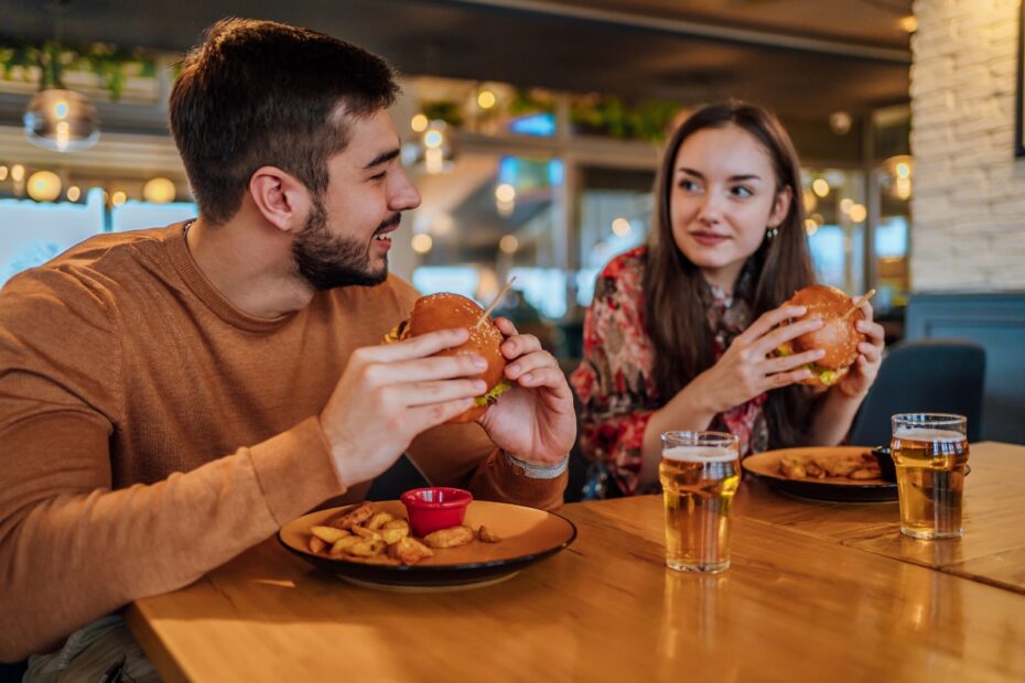 A man and a woman are seated across from each other at a wooden restaurant table, while holding burgers