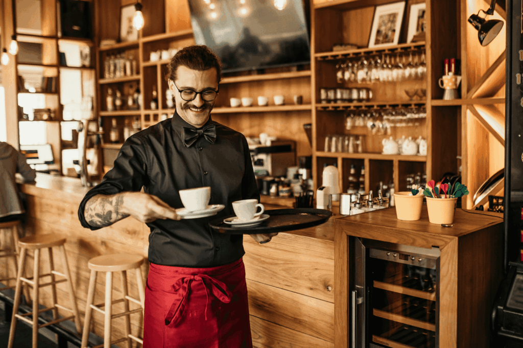 A smiling barista in a café, dressed in black with a red apron, carrying two cups of coffee