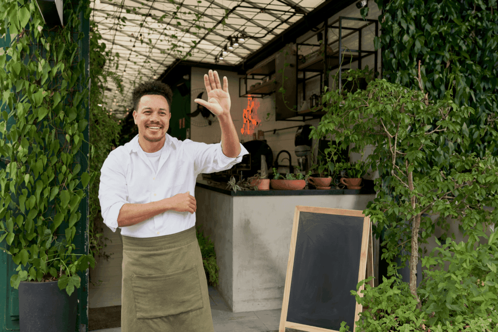 Chef in an apron waving outside a restaurant entrance.