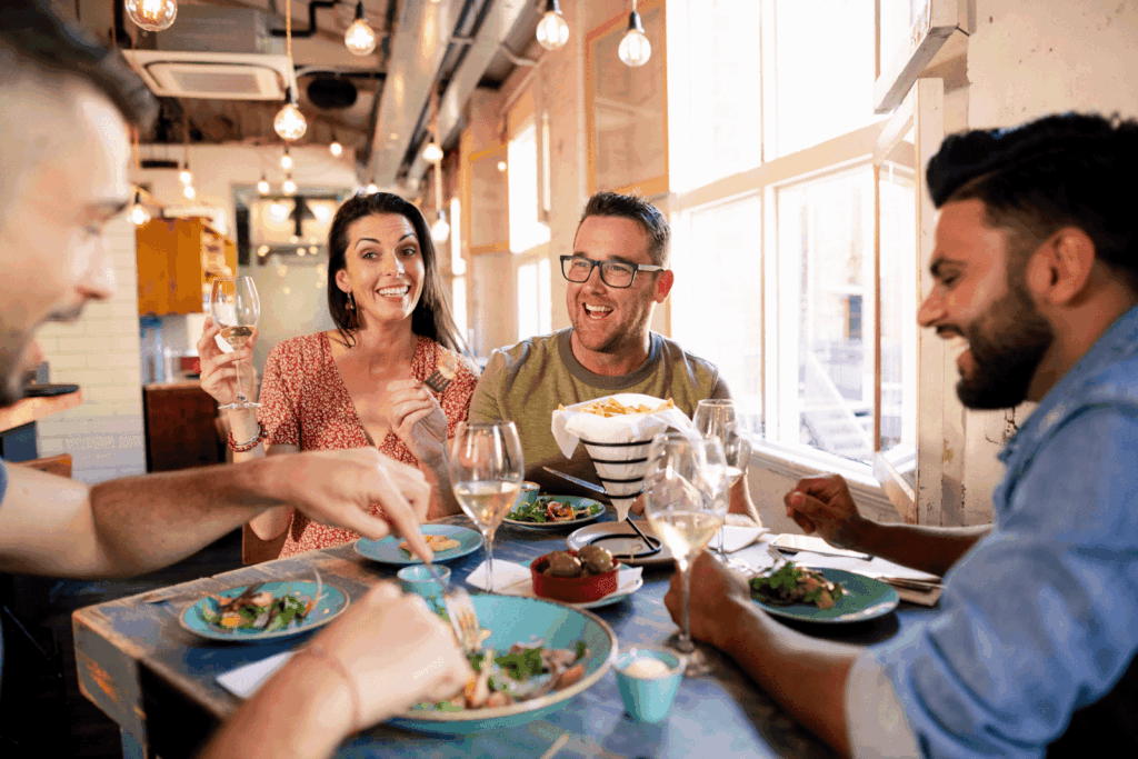 Four friends enjoy a lively meal together, smiling, in a well-lit, modern restaurant.