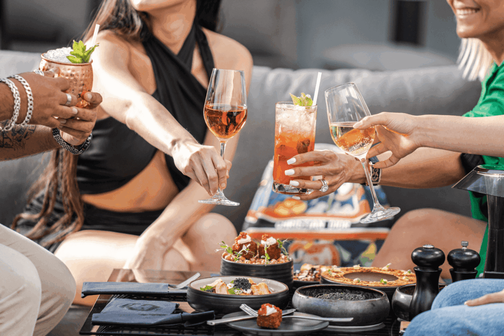 People clinking glasses over a table filled with appetizers and cocktails.