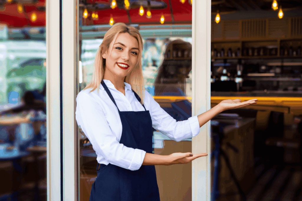 Smiling waitress standing at the entrance of a restaurant, gesturing to welcome guests.
