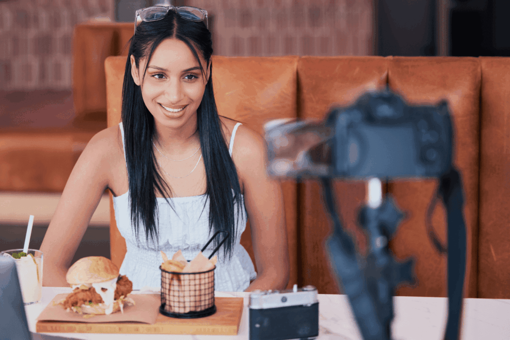 Woman sitting in a booth smiling while recording a food vlog with a camera.