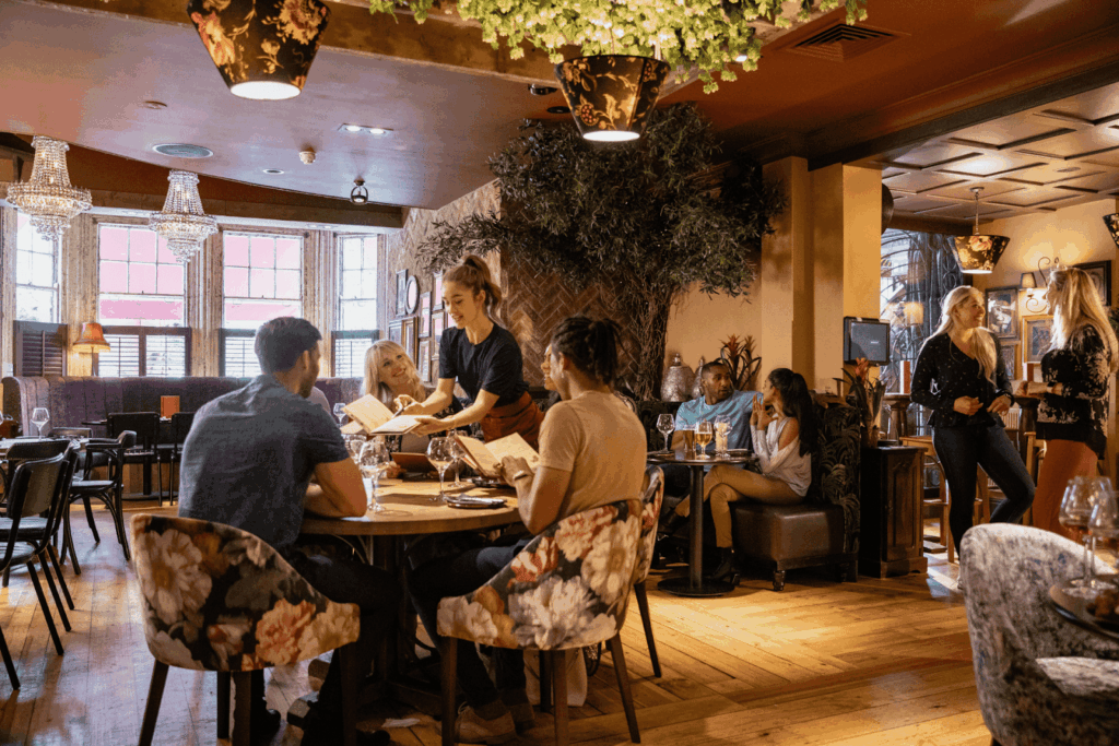 A busy restaurant interior with a server handing menus to seated guests and other people chatting around the room.