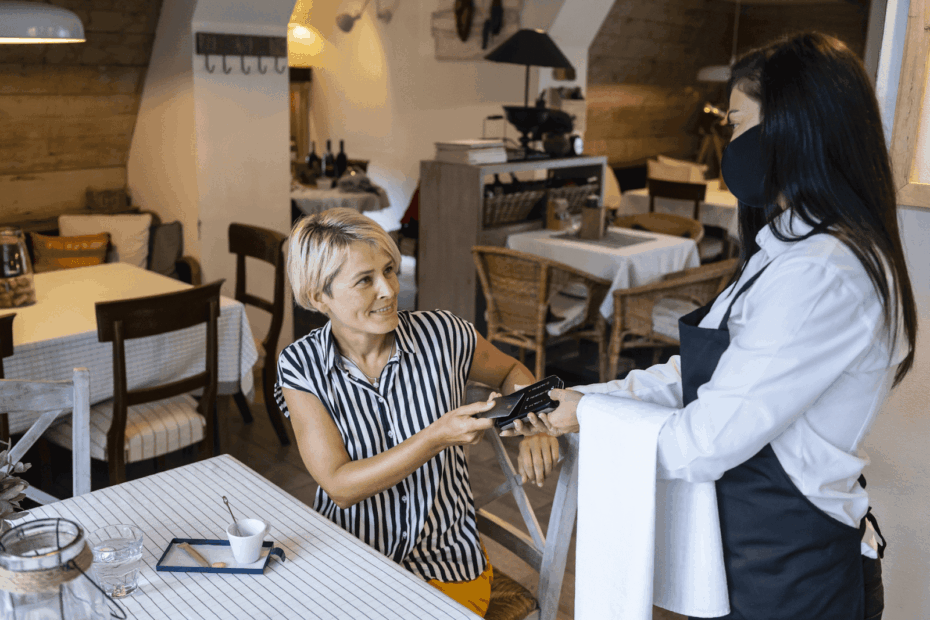 A customer paying a masked server with a card inside a cozy restaurant.
