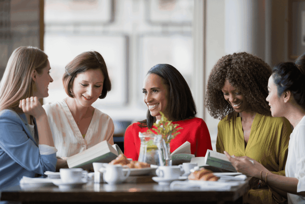 A group of women sitting at a café table, smiling and reading books together.