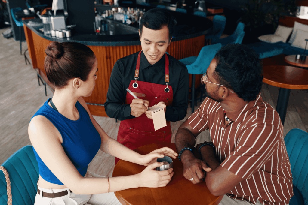 A server in a red apron taking an order from a couple seated at a café table.