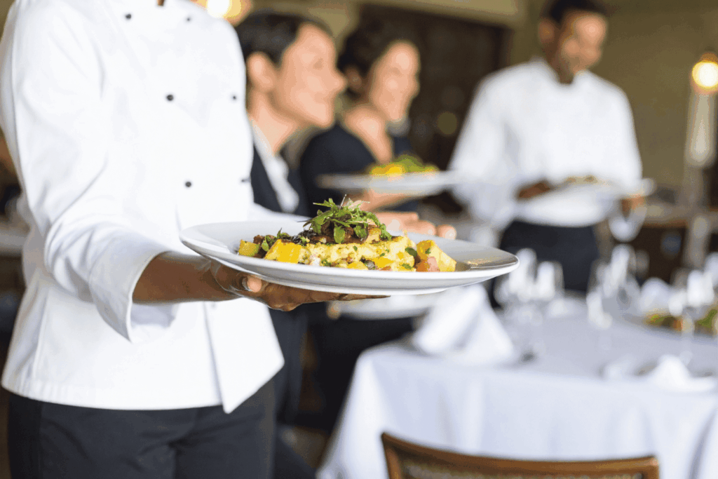 A server in a white chef coat carrying a plated dish in a busy restaurant.