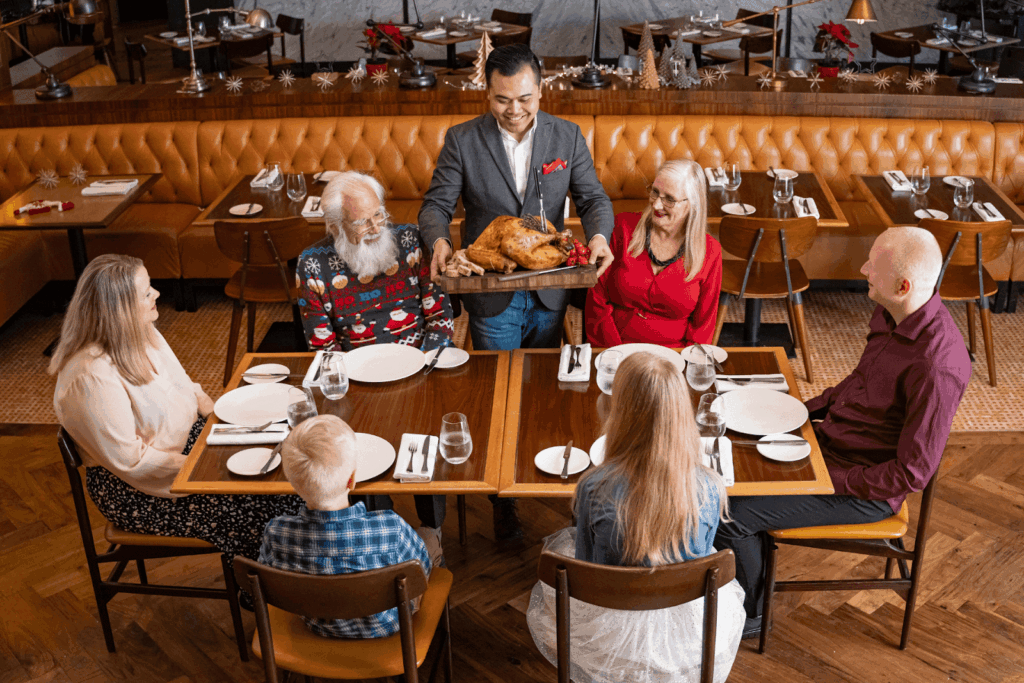 A server presenting a roasted turkey to a family seated at a holiday dinner table.