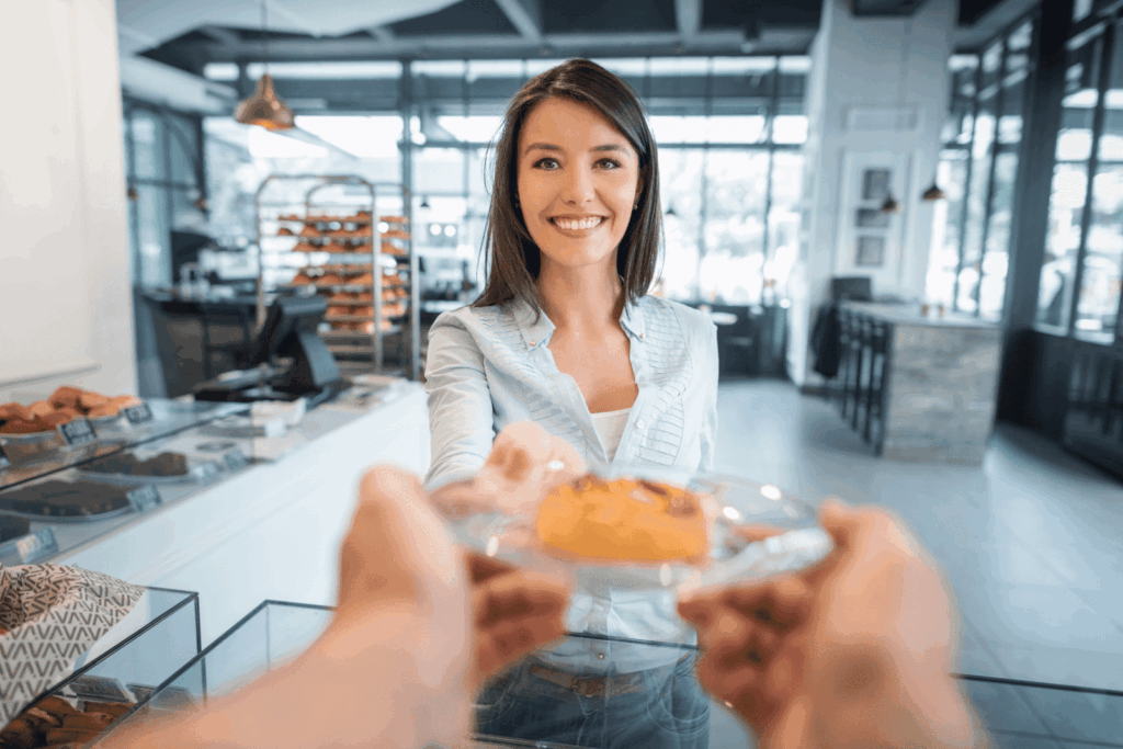 A smiling woman behind a bakery counter handing a pastry to a customer.