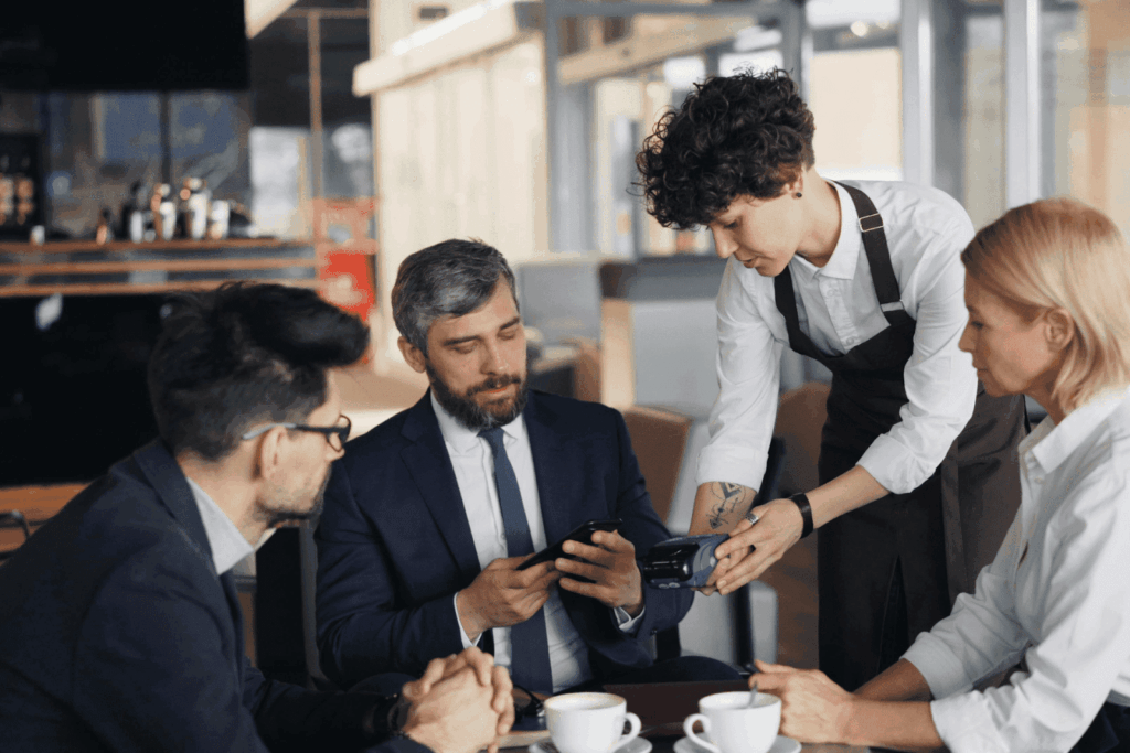 A waiter holding a card reader while a business group pays at a café table.
