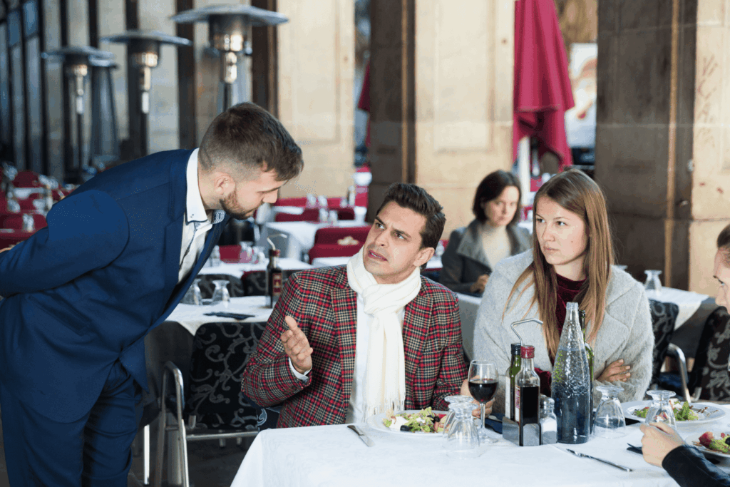 A waiter speaking with diners at an outdoor restaurant as one guest appears upset.