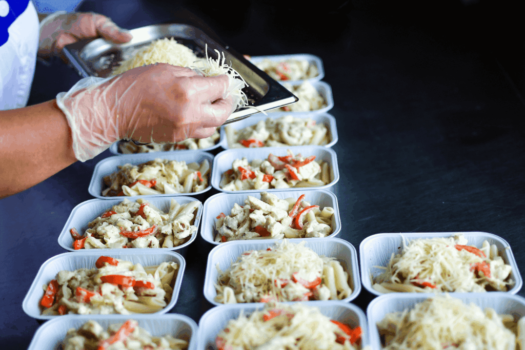 A worker preparing multiple pasta meal trays, adding shredded cheese on top.