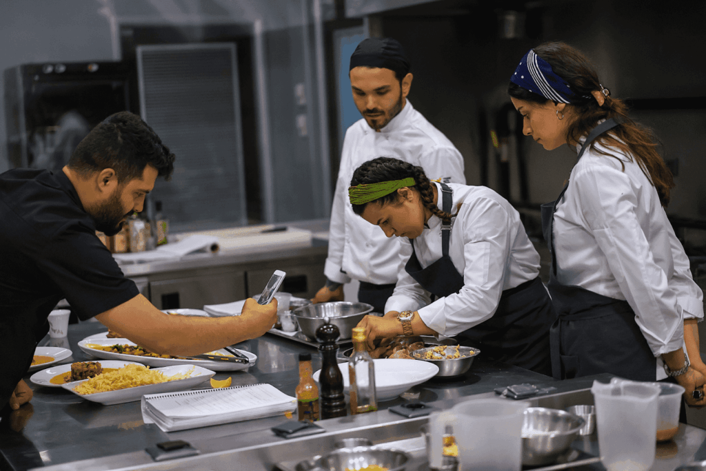 Chefs in a commercial kitchen preparing dishes while another person photographs the food.