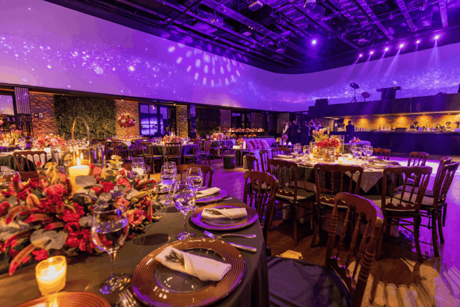Elegant event hall with decorated tables, flowers, candles, and purple ambient lighting.