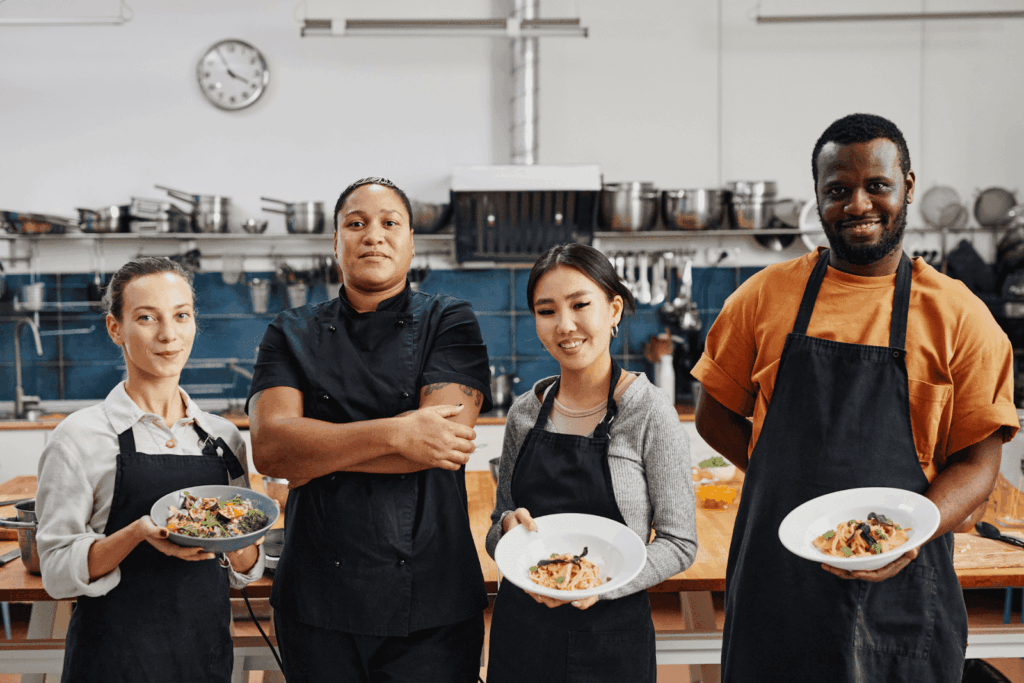 Four kitchen staff standing and holding plated dishes in a commercial kitchen.