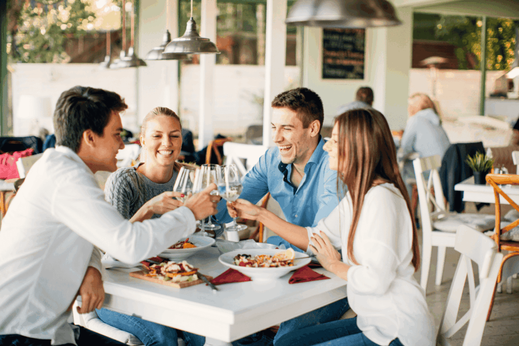 Friends clinking glasses and laughing while dining at a bright café.