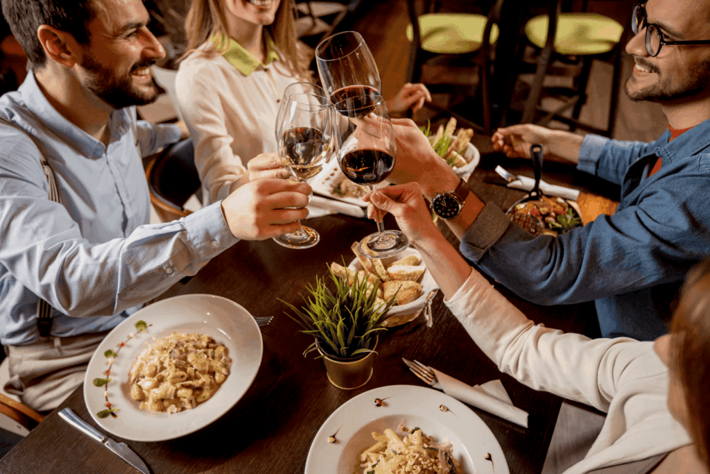 Friends clinking wine glasses over a restaurant table filled with pasta dishes.