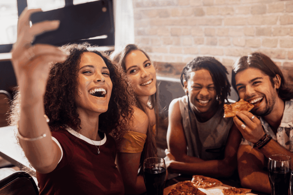  Friends laughing and taking a selfie while eating pizza together.