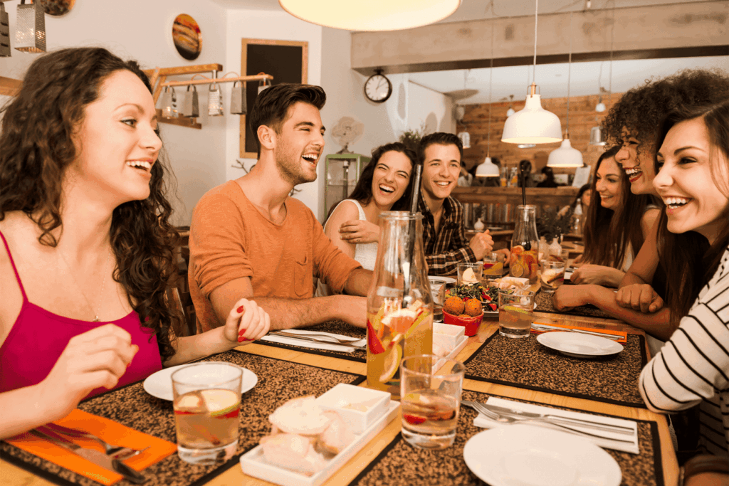  Friends laughing and talking around a long restaurant table filled with food and drinks.