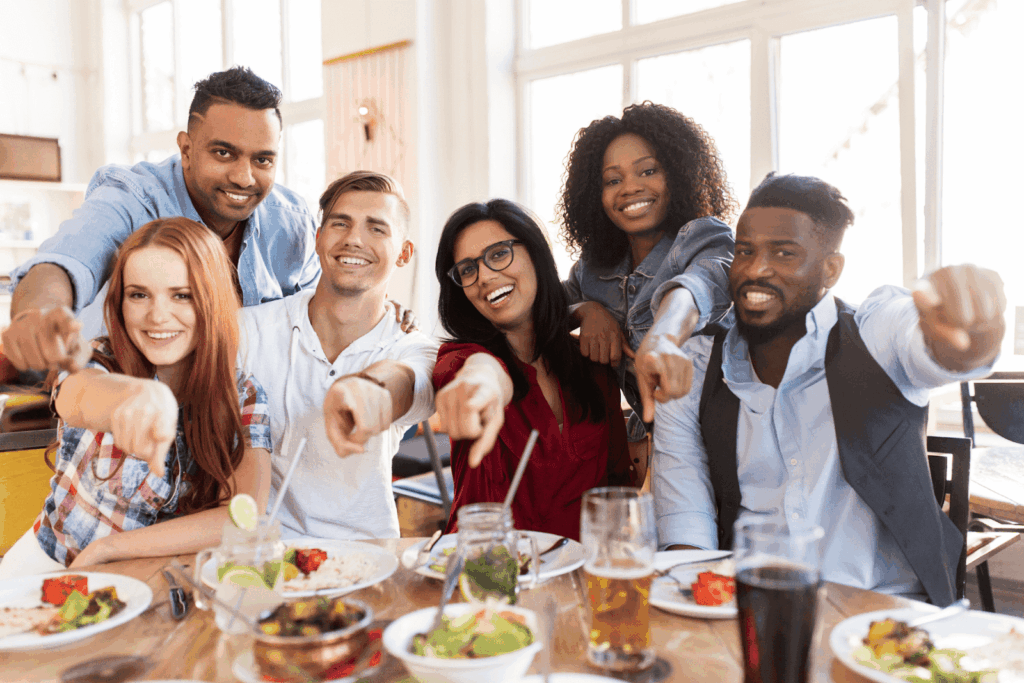 Friends sitting at a restaurant table, smiling and pointing playfully toward the camera.
