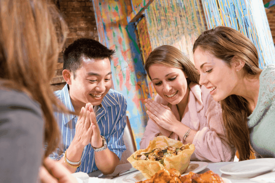 Friends smiling and looking excited as they sit around a table with food.