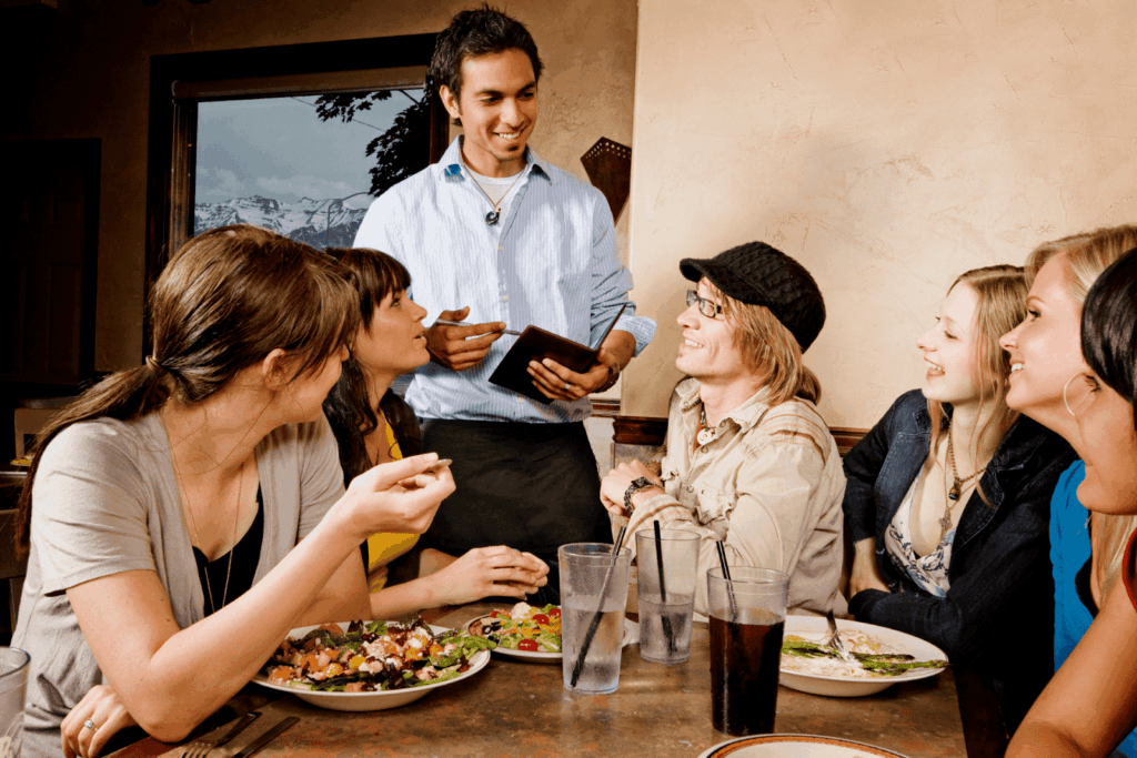 Group of friends talking with a smiling waiter while ordering food at a restaurant table.
