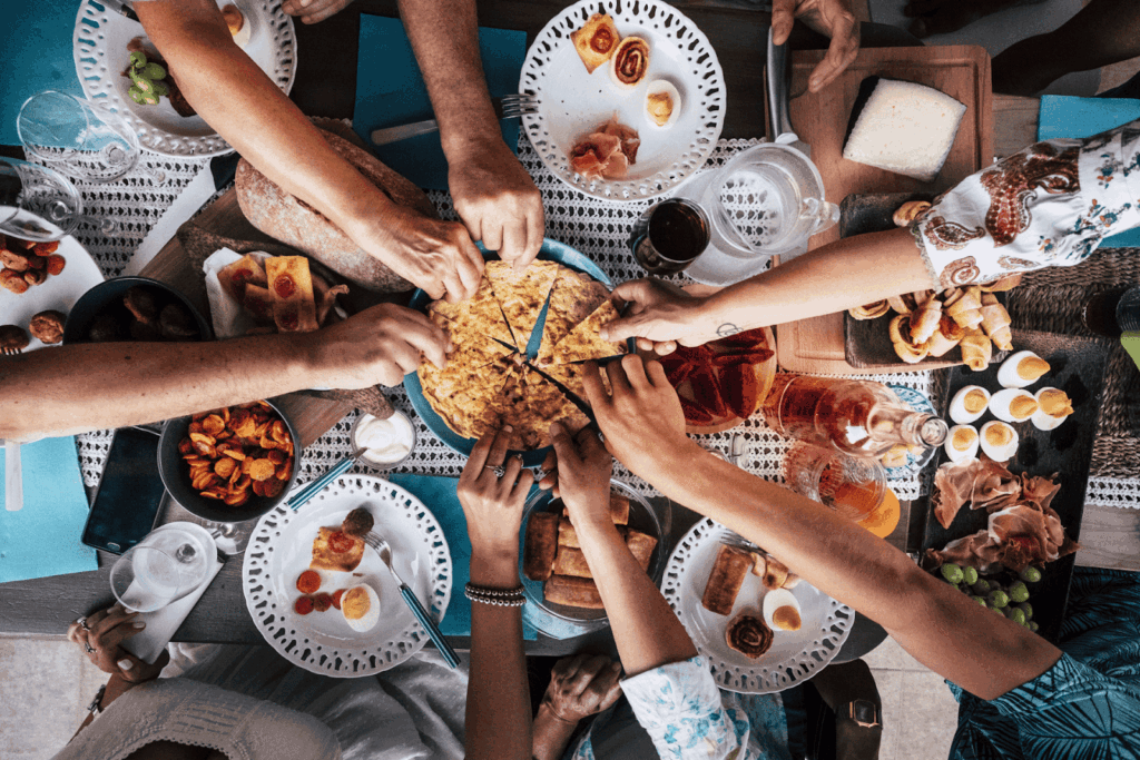 Group of people reaching in to share a large dish at a crowded table of assorted foods.