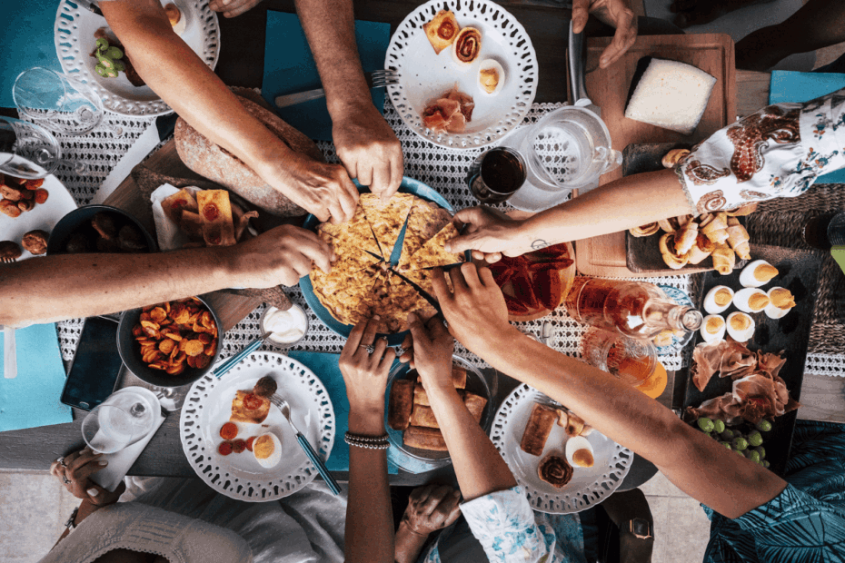 Group of people reaching in to share a large dish at a crowded table of assorted foods.