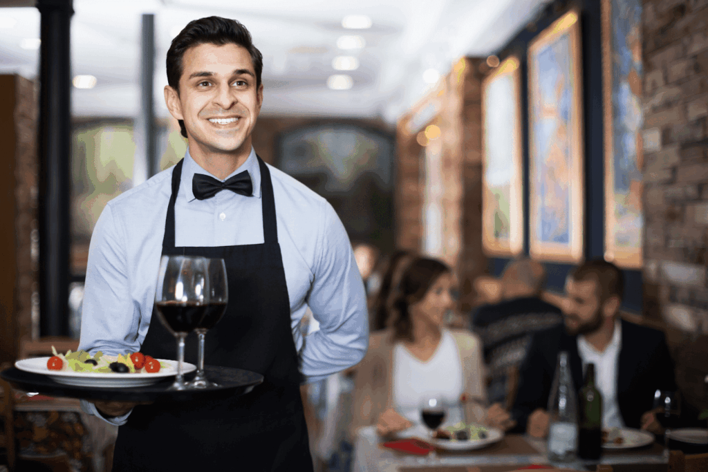 Smiling waiter carrying a tray with wine and a salad in a busy restaurant.