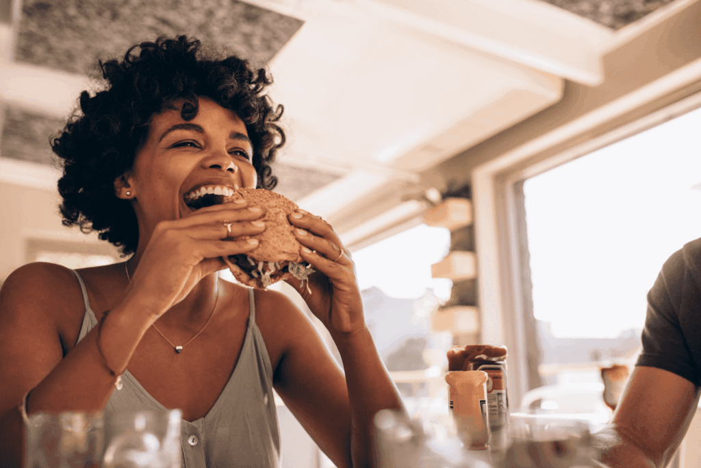 Smiling woman taking a big bite of a burger at a bright, casual restaurant.