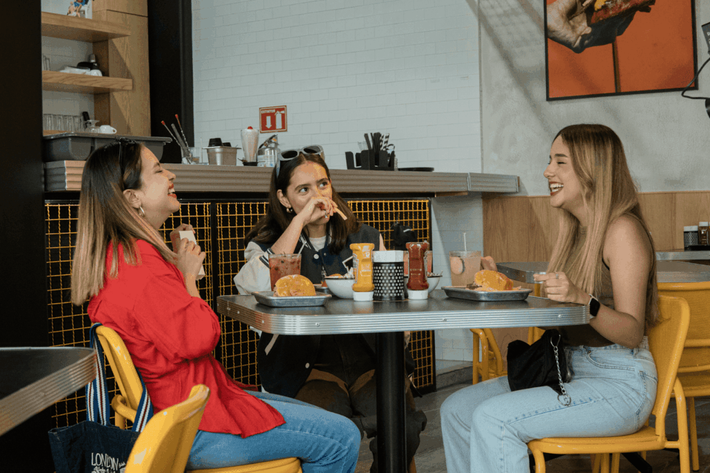 Three friends sitting at a casual restaurant table, laughing and eating burgers.