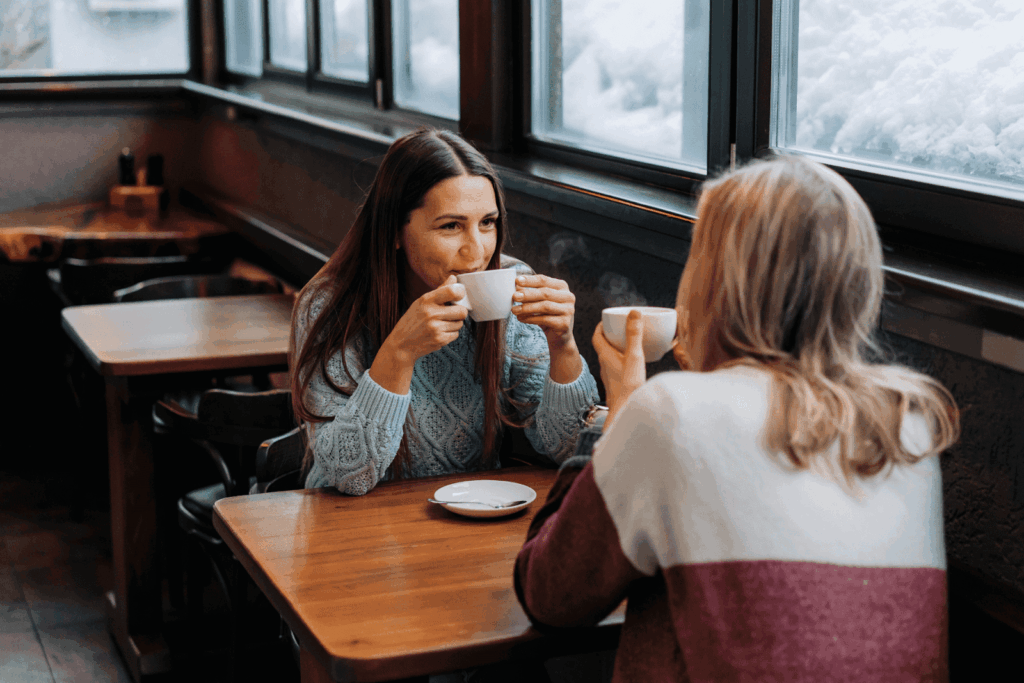 Two women sitting in a café by a snowy window, drinking hot beverages and chatting.