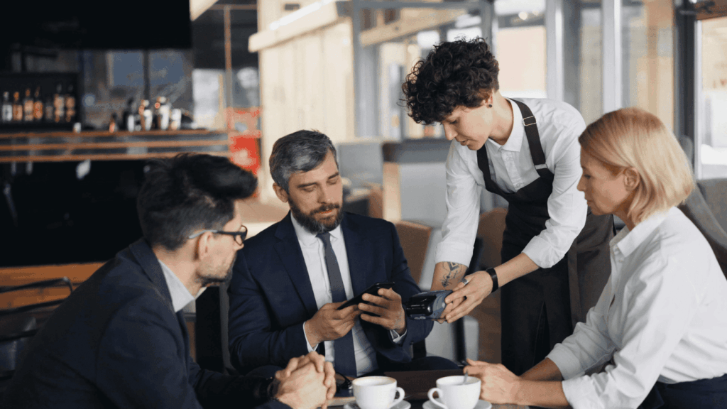 Waiter holding a payment terminal while a man at a table taps his phone to pay during a business meeting.