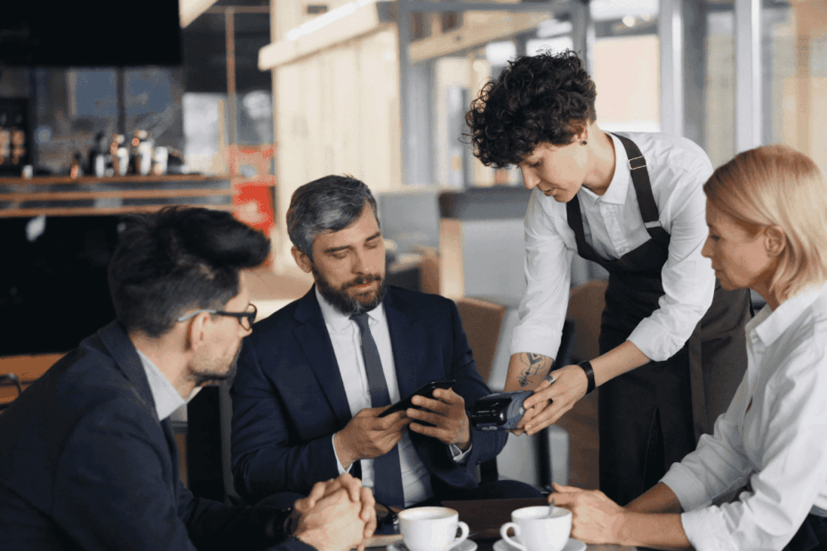 Waiter holding a payment terminal while a man at a table taps his phone to pay during a business meeting.