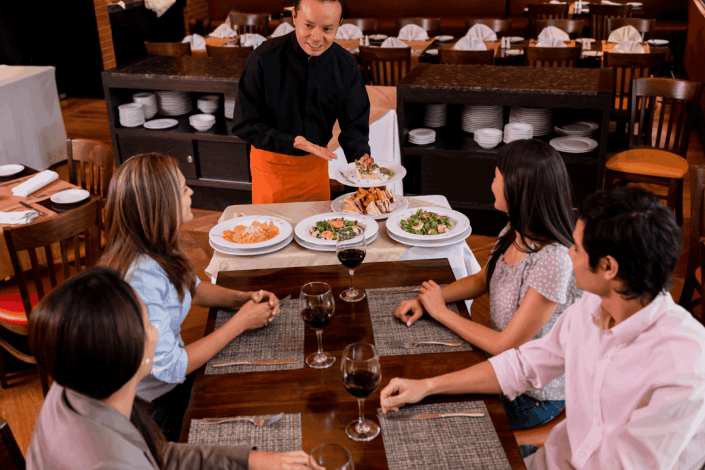 Waiter presenting several plated dishes to a group of diners at a restaurant table.
